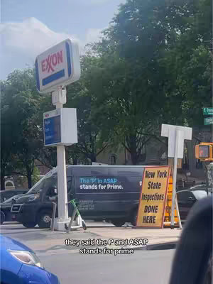 An outdoor scene at a gas station. The focus is on the Exxon sign, a van with text, and a sign that says 'New York State Inspections Done Here'. In the foreground is part of a blue car, and a person's point of view while filming is captured through the car.