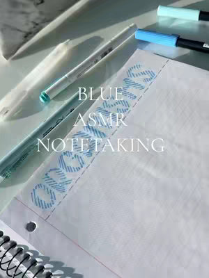 A still life captures a collection of school or office supplies arranged on a light, possibly marble-like surface with soft lighting. A spiral notebook rests at the bottom, with a few blank white pages visible. Above, several pens and markers in various shades of blue create a visual harmony. Text overlays in a light color on top of the notebook. Overall, the image has a clean, well-lit aesthetic.