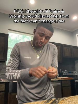 A man in a gray shirt stands in a kitchen, looking down and slightly to the side. Text overlays the image: POV: I thought a lil Praise & Worship would distract her from the fact I can't change a tire.