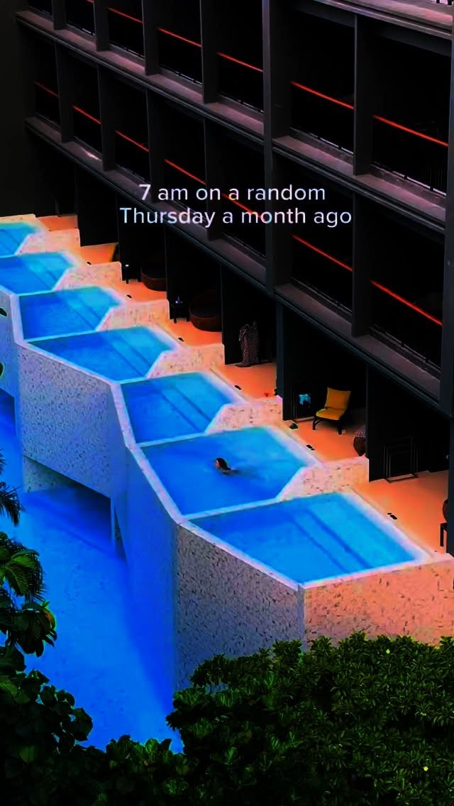 An aerial view showing a luxurious hotel with a series of interconnected swimming pools cascading down the side of the building. The pools are a bright blue, with a person swimming in one. The architecture is modern, and the image is partially obscured by green foliage in the bottom and sides. Text overlay states the time and date of the photo.