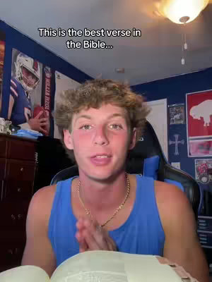 A young man with curly brown hair is centered in the frame, wearing a blue tank top and holding an open book. Text overlays the image: "This is the best verse in the Bible..." In the background, a room is visible with posters, a fan, and other decor.