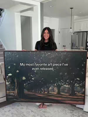 A woman stands in front of a large framed painting of a tree-lined road. The painting depicts a serene landscape scene in muted colors. The woman is smiling, and the scene is lit by natural light.
