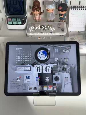 The image showcases an iPad displaying a customized home screen against a backdrop of decorative items. The iPad, centrally positioned, sits on a clear stand, its screen filled with app icons, widgets, and a stylized wallpaper featuring a calendar, digital clock, and other design elements. Behind the iPad, a shelf holds various decorative items, including figurines of cartoon characters, and a calendar. On top of the shelf, a small keyboard and a case for the iPad are visible. The overall aesthetic suggests a focus on organization, personalization, and a touch of cuteness.