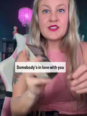 A woman with long blonde hair and blue eyes is looking at the camera while holding a deck of cards, with the text overlayed on the image saying, "Somebody's in love with you". The woman is wearing a pink top, and the background includes a lamp, and a chair.