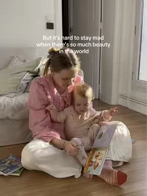 A woman and a young child are seated on a wooden floor, lit by natural light from a window. The woman, with blonde hair pulled back, wears a pink blouse and white pants. The child, wearing a light pink outfit and a pink bow, sits on the woman's lap, both looking at a colorful book. The background includes a neutral-colored wall, a window, and a portion of a white couch.
