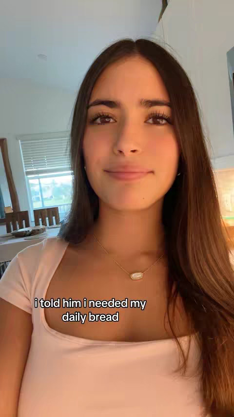 A young woman with long brown hair is centered in the frame, looking slightly off-camera with a small smile. She wears a light pink top and a delicate gold necklace. The background features a partially visible interior, suggesting a home environment with glimpses of furniture and decor.