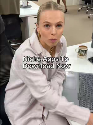 A woman with blonde hair is seated at a desk in an office setting. She is wearing a light-colored, collared shirt and gold hoop earrings. The image has text overlayed, with a laptop visible on the desk and a coffee mug in the background.