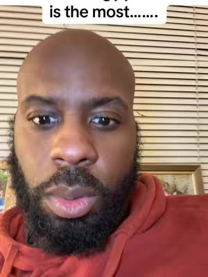 A close-up shot of a Black man with a shaved head and a full beard. He is wearing a red hoodie. The background consists of horizontal blinds and a framed picture. The man's expression is neutral, as if he's about to speak.