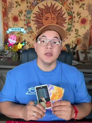 A man wearing glasses and a hat is holding up two tarot cards. He is positioned in front of a tapestry with a sun design. A banner that says 'PISCES' floats near his head. The man is wearing glasses and a blue shirt.
