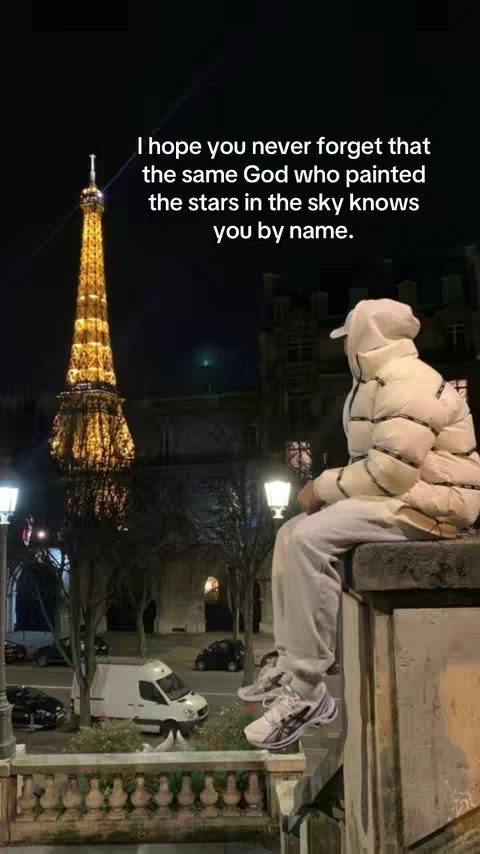 A nighttime scene with the Eiffel Tower lit up in gold, a person wearing a white puffer jacket and a hood is sitting on the ledge. The overall tone of the image is contemplative and serene.