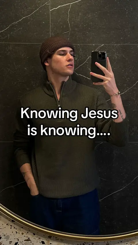 A man is taking a mirror selfie. He is wearing a brown beanie, a green zip-up sweater, and dark blue jeans. The background is a dark marble wall. Text overlayed on the image reads, "Knowing Jesus is knowing...."