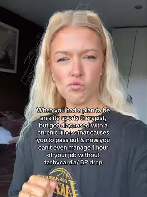 A medium shot of a young woman with long blonde hair, making a disgruntled face. The background is blurred, suggesting an indoor setting. She appears to be in front of a door or dark background.