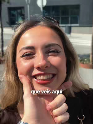 A woman with blonde hair smiles widely, her hand holding another hand that is pointing towards her face. The background is blurred, showing elements of an urban environment.