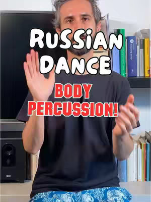 A man in a black t-shirt with the overlay text 'RUSSIAN DANCE BODY PERCUSSION!' stands in front of a television and bookshelves. He is gesturing with his hands as if performing a body percussion routine.