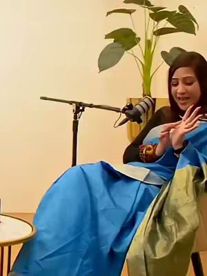 A woman sits in a brightly lit space, gesturing with both hands while looking towards the right. A microphone is positioned in front of her, and a large plant is in the background. She is wearing a blue and gold outfit, sitting on a light-colored chair.