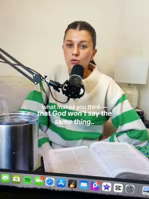 A woman is centered in the frame. She's wearing a sweater with green and white stripes. A microphone is positioned in front of her. On the table, there is an open book. The background consists of a white wall, a lamp, and other objects. A text overlay near the bottom of the screen.