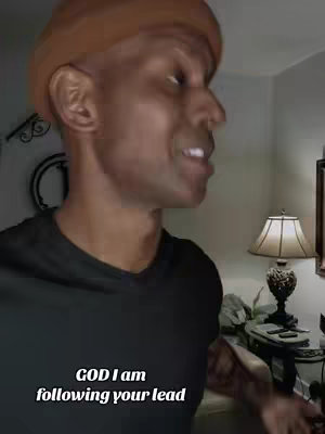 A man with a brown beanie is speaking, likely to or at the camera, with text overlayed at the bottom against a backdrop of what appears to be a living room setup. The frame is a medium shot capturing his face and upper body, focusing on his expression and the surrounding environment. Soft lighting and a lampshade in the background contribute to a warm atmosphere.