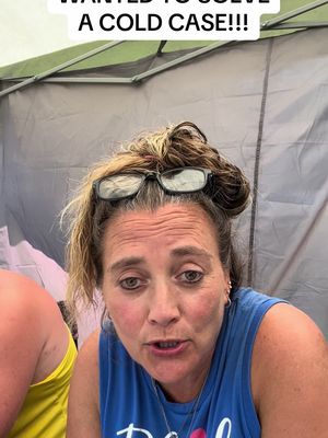 A woman with blonde hair and glasses on her head is looking directly at the camera with a concerned expression. She is wearing a blue tank top. The background is a light-colored canvas or tent material.