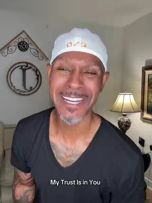 A smiling man is centered in the frame, wearing a black v-neck t-shirt and a white cap with multicolored rectangles on it. His face is filled with a beaming smile, showing his teeth and a short salt and pepper beard. Behind him, a cream-colored wall has a decorative wooden piece and a large circular object with the letter 'T' on it. To the side, a table lamp with a tan lampshade provides soft lighting.
