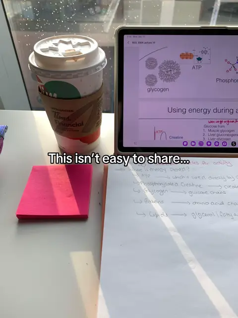 A top-down view of a desk setup with a laptop displaying a biology diagram, a coffee cup, a pink post-it note, and a handwritten note sheet. The laptop screen shows colorful illustrations and text related to a science topic. The background includes a window with diffused natural light.