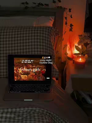 A laptop displaying a "Gilmore Girls" scene rests on a bed covered with a checkered blanket. A lit candle and a small plant on a nightstand illuminate a dimly lit room. Additional decor includes a vine of green leaves.