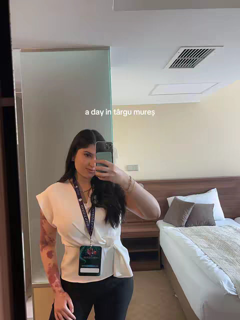 A woman is taking a selfie in a mirror, with a blurred background showing a room with a bed. She is wearing a white top and black pants, holding a phone to take the picture. There's text overlayed on the image.