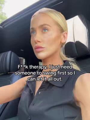 A woman sits in the driver's seat of a car, looking sideways with a serious expression. She has blonde hair pulled back, and her skin is fair with makeup. The interior of the car is visible, and text is overlaid on the image.