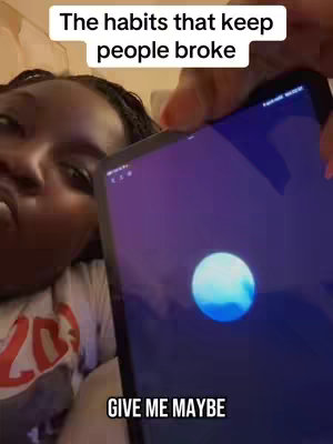 A woman with dark skin is lying in bed and holding a tablet, the screen of which is mostly obscured. Two lines of white text are in a white rectangular box, "The habits that keep people broke" and "GIVE ME MAYBE" in smaller text are in the lower part of the image.