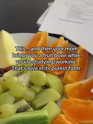 A close-up shot of a fruit bowl filled with various fruits such as kiwi, oranges, grapes set against a blurry background. The text overlay reads, "This... and then your mom brings you a fruit bowl while you're studying/working. That's love in its purest form."