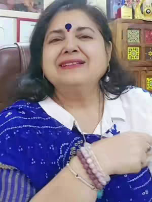A woman with dark hair and a bindi smiles and looks towards the camera. She wears a blue and white patterned shirt and jewelry. The background includes a wooden cabinet and framed pictures.