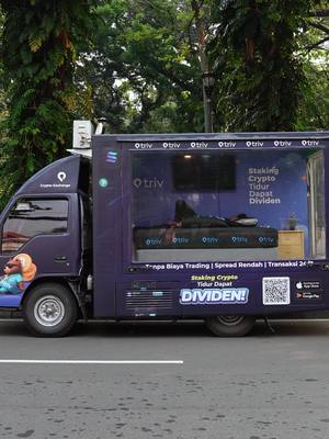A truck in an urban setting with large transparent windows showcasing a bed and various crypto-related branding. The truck is dark purple with prominent 'trlv' and 'CryptoWave' logos. Text overlays promote 'Staking Crypto' and 'Dividen'. The background is somewhat blurred with trees.