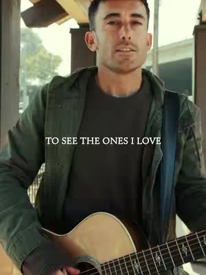 A medium shot of a man playing an acoustic guitar outdoors. He's wearing a dark green jacket over a gray shirt. Text overlayed on the image says, "TO SEE THE ONES I LOVE". The background is blurred, suggesting an outdoor setting.