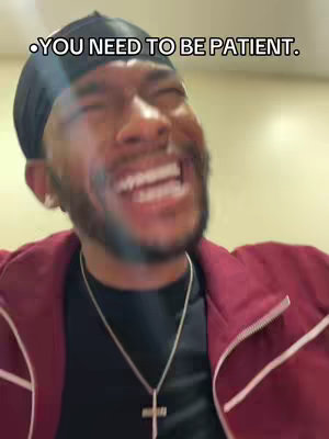 A man with a beard is shown smiling widely, with his head tilted slightly towards the right. He has a head covering, a dark shirt, and a maroon jacket. A silver-colored cross necklace is visible. Text overlayed at the top of the image says, 'YOU NEED TO BE PATIENT.'