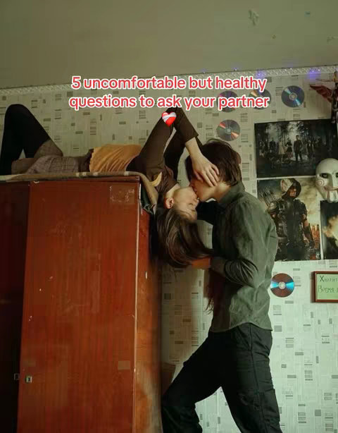 A couple is shown kissing in a close embrace. The woman is lying upside down on top of a wooden cabinet, and the man is standing next to her, holding her. Text on the wall suggests decorative elements in the background.
