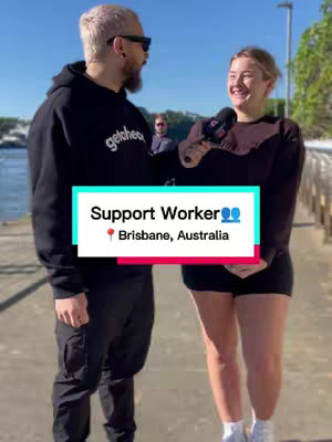 A shot of two people, a man and a woman, standing on a paved pathway next to a body of water. The man on the left is wearing a black hoodie, and the woman on the right is holding a microphone and smiling. In the background are trees and a building, as well as text overlays.