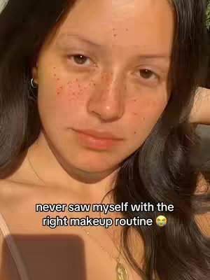 A selfie-style shot showing a woman's face, split down the middle. The left side of her face shows no makeup, natural skin with freckles, and the right side has makeup applied, potentially with a fuller look. The woman has dark, wavy hair and a neutral expression. Text overlay at the bottom reads a message. The overall composition is well-lit with soft, natural lighting.