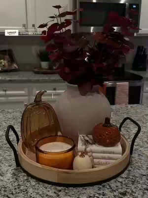 A shot of a kitchen countertop styled for fall. A wooden tray holds a candle, glass pumpkin, ceramic pumpkin, stacks of books, and a vase with leafy stems. The background shows a kitchen with stainless steel appliances.