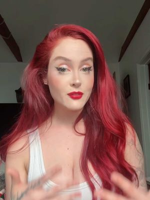 A medium shot of a woman with vibrant red hair, styled with loose waves. She is wearing a white tank top and has full makeup with red lipstick and winged eyeliner. Her arms are raised. The background is a room interior.