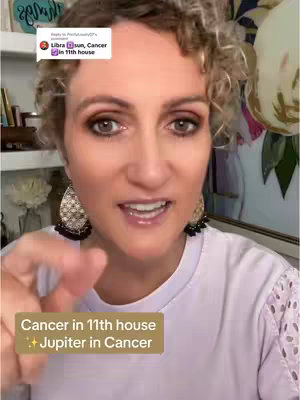 A woman with curly blonde hair, wearing gold hoop earrings, and a white top is shown from the chest up. She is speaking and gesturing with her right index finger is pointing to the space between her face and the camera. There is text on a yellow rectangle in the bottom third of the frame saying "Cancer in 11th house + Jupiter in Cancer". A comment from a user is in the top-left.