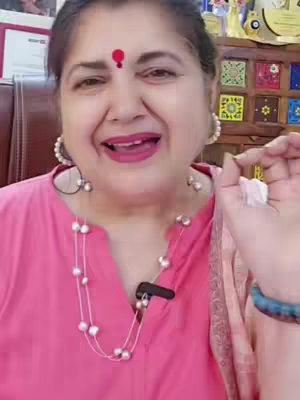 The image features a woman with fair skin, wearing a pink top and a pearl necklace. She has a red dot on her forehead and is smiling with her mouth open, possibly speaking. Her dark hair is pulled back, and she's holding something white in her right hand. The background includes a blurred room interior with wooden furniture.