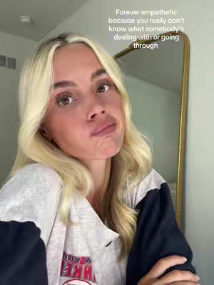 A blonde woman with a slightly upward turned pout looks directly forward. Her left hand angles up and grips the edge of the frame. She wears a light gray top over a dark blue sweatshirt. Behind her is a gold-framed mirror.