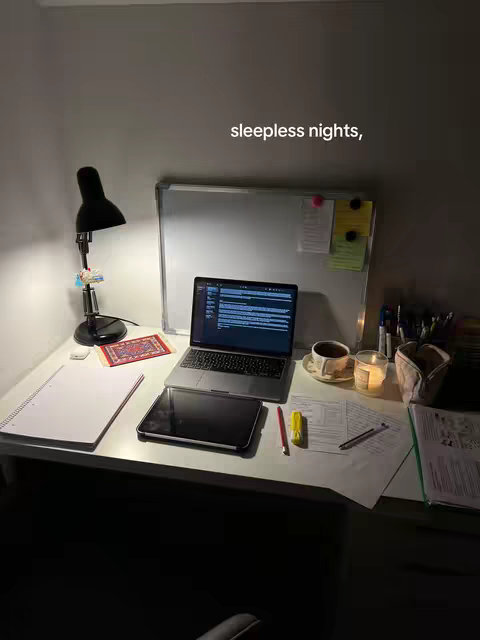 A dimly lit desk with a laptop displaying a document. A tablet and a notepad are also on the desk. There is a mug, a candle, documents, pens, and a lamp visible. The setting appears to be a workspace or study area. 