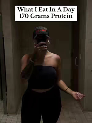 A person is standing in a hallway with a cap in hand and phone covering the face. Text overlay says, "What I Eat In A Day 170 Grams Protein."