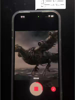 A phone displaying a video of a mechanical bird-like creature. The phone is held up against a black surface. A text bubble with a series of marks or symbols is in the top right corner.