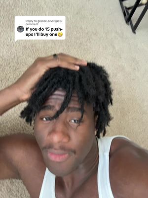 A selfie-style shot featuring a young man. He is lying down, with his head slightly elevated. His body is partially visible. He is wearing a white tank top. His face is the main focal point. He has dark hair styled back and a mustache. He is looking towards the camera with a slightly inquisitive expression. A comment from an online platform is visible in the background, partially visible on the left side of the frame.