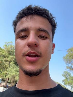 A close-up shot of a young man with a short beard and mustache, looking directly at the camera. He has short, curly dark hair and is wearing a black shirt. The background is a bright blue sky with some trees visible on the sides.