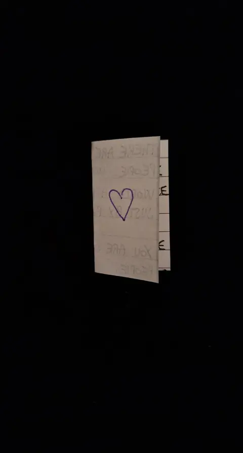 A folded piece of paper, possibly a card, is the central focus, set against a stark black background. A hand-drawn purple heart is visible on the front of the card, which looks roughly made. Text is partially visible on the folded section. The composition is simple and isolated, which suggests a message of intimacy. The overall look is plain and lacks any color apart from the heart.