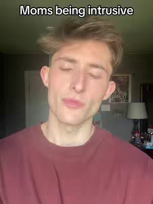 A selfie-style video cover with a young man as the main subject. He's in a room, possibly his home, with a blurred background that includes a door, a lamp, and a hint of decor. The man's expression is neutral. The text overlay reads, "Moms being intrusive."