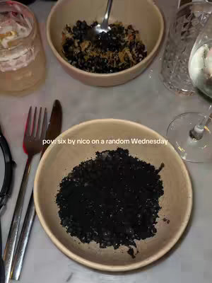 A slightly overhead shot of a table setting at a restaurant. Two bowls are visible, one in the upper half filled with a dark, textured food mixture and the other in the bottom half, prominently featuring a mound of black food resembling caviar. Silverware, including a fork and knife, are positioned on the left side of the frame in alignment with a clear glass of water, a cocktail glass, and a partially seen wine glass on the right side.