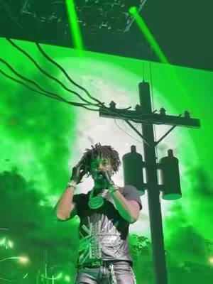 The cover image features a male performer on stage, illuminated by vibrant green lighting. He is dressed in a black outfit with silver accents, including a vest and jewelry. The background includes a stylized image of a utility pole and a moon, also in shades of green. The overall composition is dynamic, capturing the energy of a live performance.